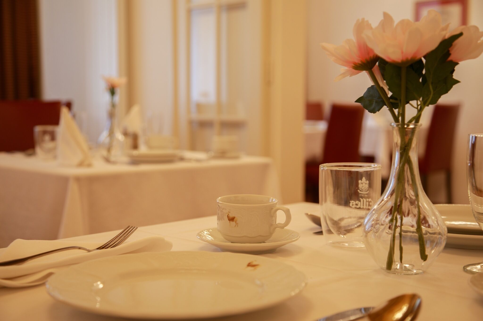 Une table à manger recouverte d’une nappe blanche, une tasse blanche ornée d’un logo représentant un cheval, un vase transparent rempli de fleurs et une cuillère.