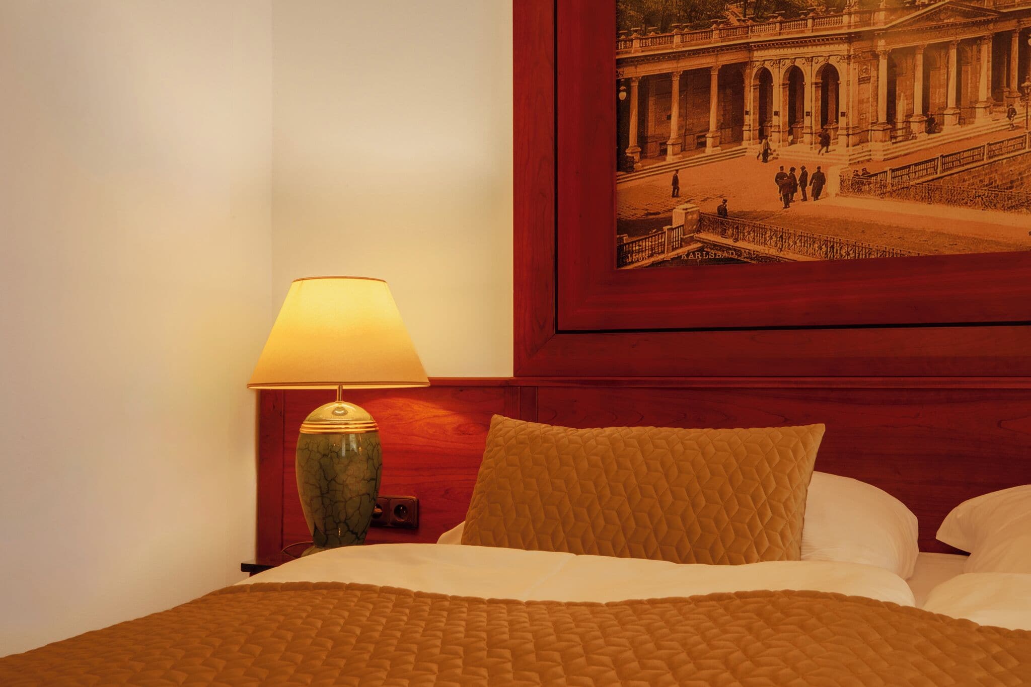 A hotel room with a bed, a lamp, a framed picture of a building, and a wooden headboard.