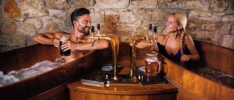 A couple in a wooden bathtub with beer taps, set against a stone wall.