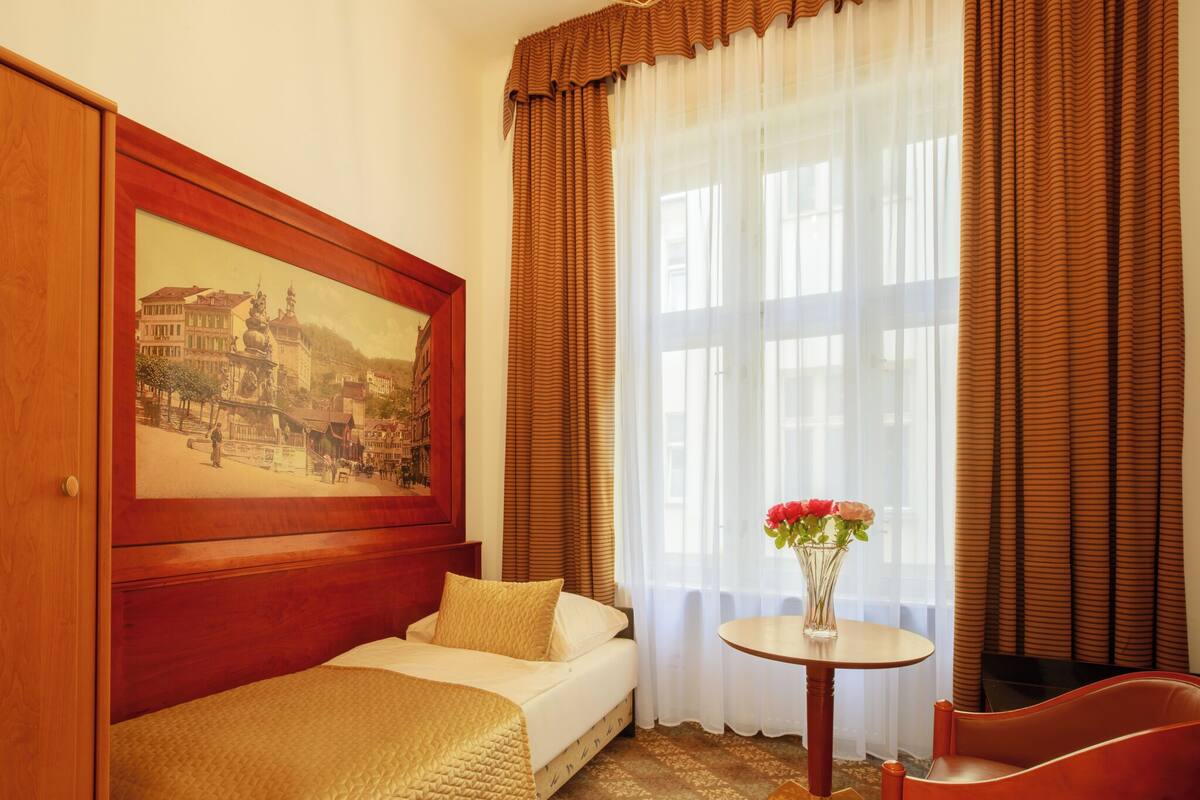 Habitación de hotel con cama, una mesita con un florero, una silla y un ventanal con cortinas.