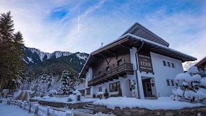 Ein zweistöckiges Alpenhotel mit Balkon, umgeben von schneebedeckten Bäumen und Bergen.
