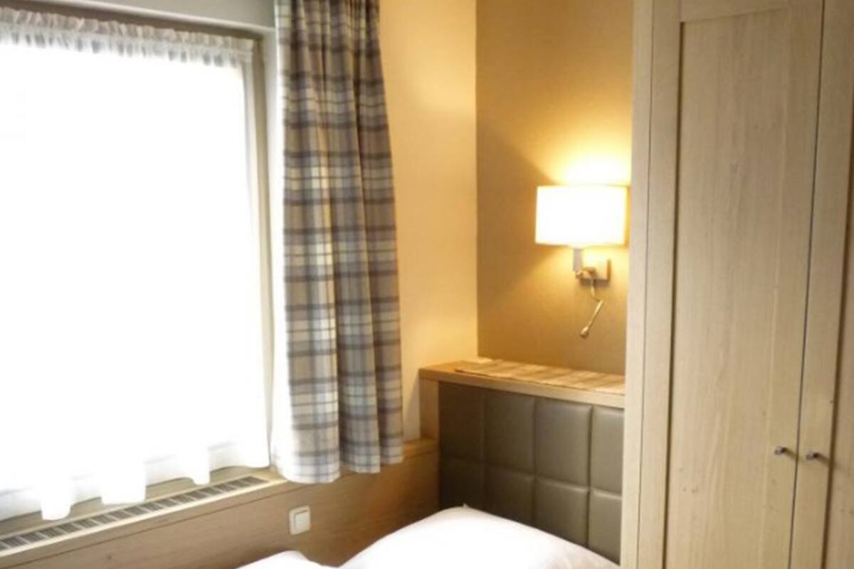 A single bed with a headboard, a window with a plaid curtain, a bedside lamp, and a wooden wardrobe.