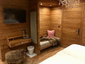 A wooden room with a bed, a TV mounted on the wall, a small wooden shelf, and a dog.