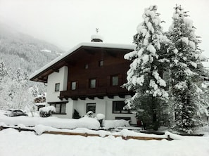 A two-story chalet with a sloped roof, surrounded by snow-covered trees and a mountainous backdrop.
