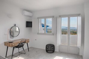 A room with a wooden desk, a black chair, a round mirror, and a television on the wall. There are two windows with white frames and a view of the landscape. - NK Luxury Villas (Naxos)
