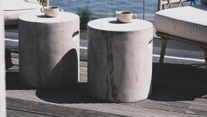 A modern outdoor seating area with wicker chairs and concrete tables, overlooking a body of water and a rocky shoreline. - Skärgårdshotellet Hönö (Hono)