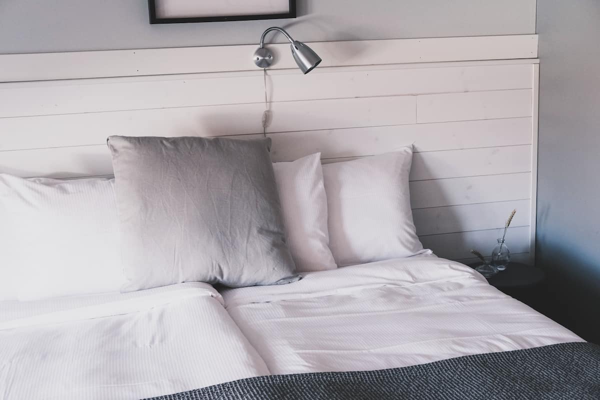 A neatly made bed with white linens and two pillows, a bedside lamp, and a small vase with dried plants on a nightstand.