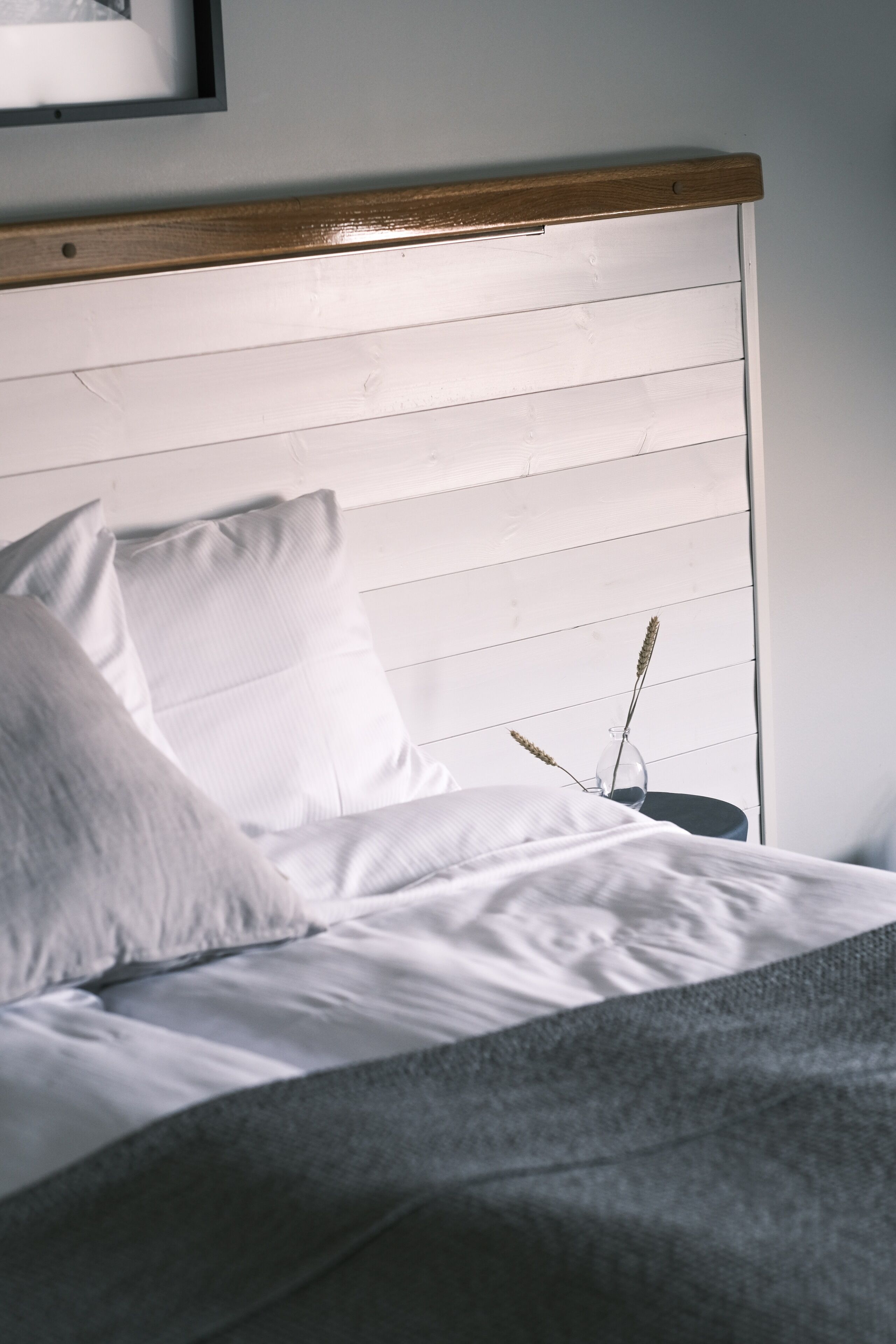 A neatly made bed with white linens and pillows, a wooden headboard, and a small table with a vase of dried plants.