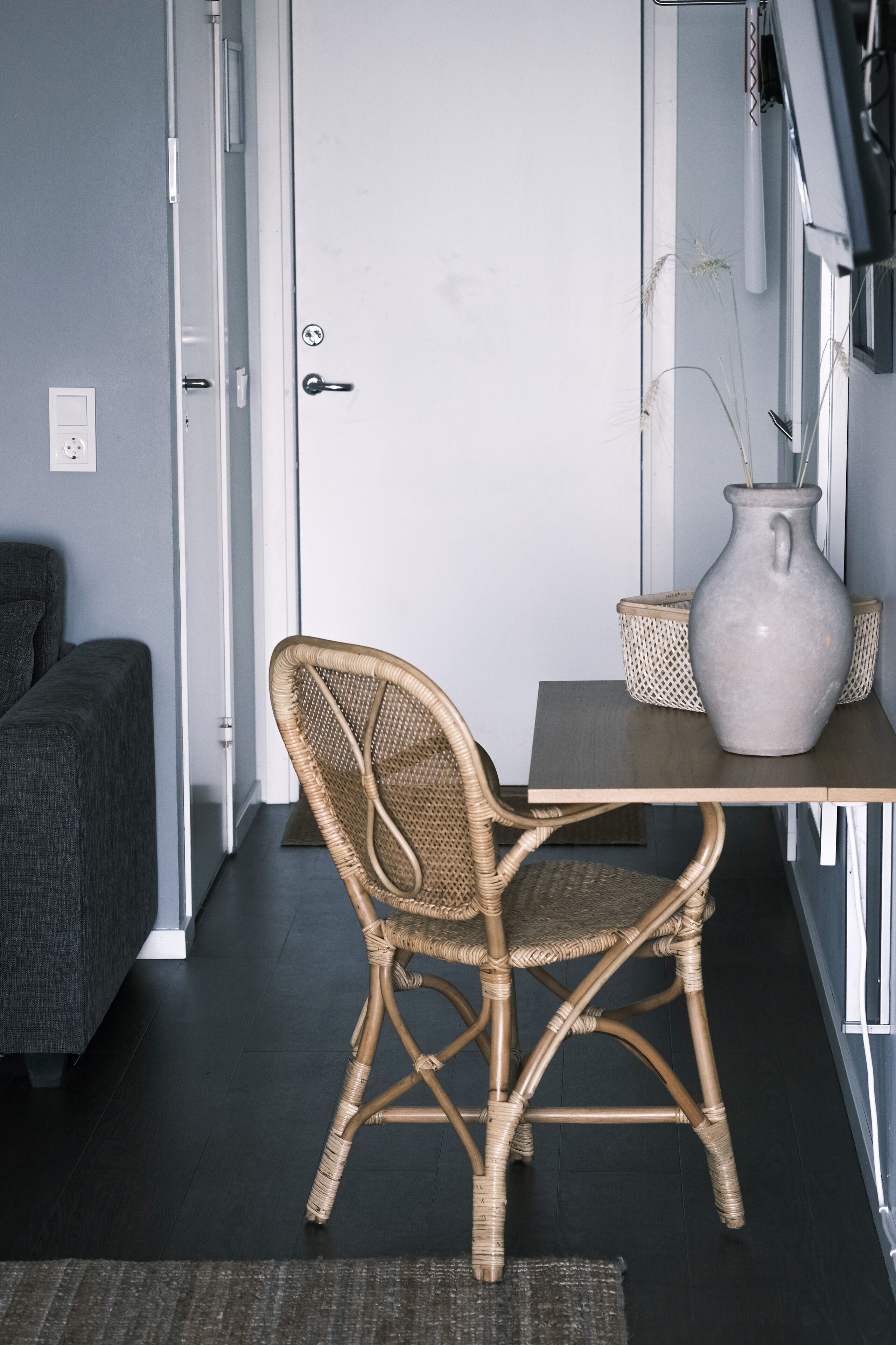 Una habitación con una silla de mimbre, una mesita, un jarrón y un sofá gris.