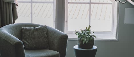 A room with a sloped ceiling, a window with a view of a bamboo roof, a grey armchair, and a potted plant on a pedestal.