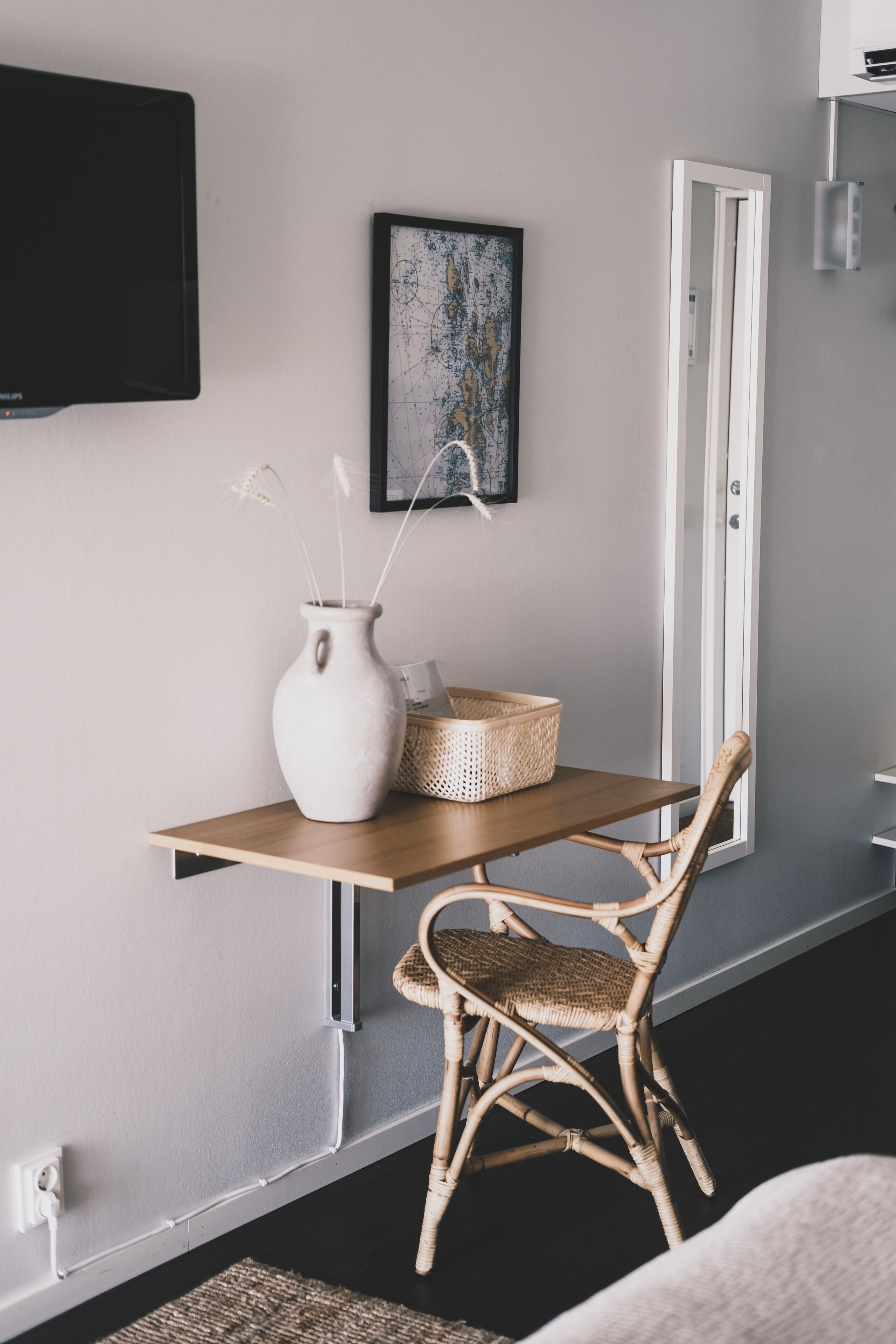 A room with a wooden desk, a wicker chair, a white vase with dried plants, a framed abstract painting, and a flat-screen TV mounted on the wall.