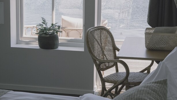A room with a large window overlooking a rocky coastline. There is a bed with white bedding, a wicker chair, and a small table with a basket. - Skärgårdshotellet Hönö (Hono)