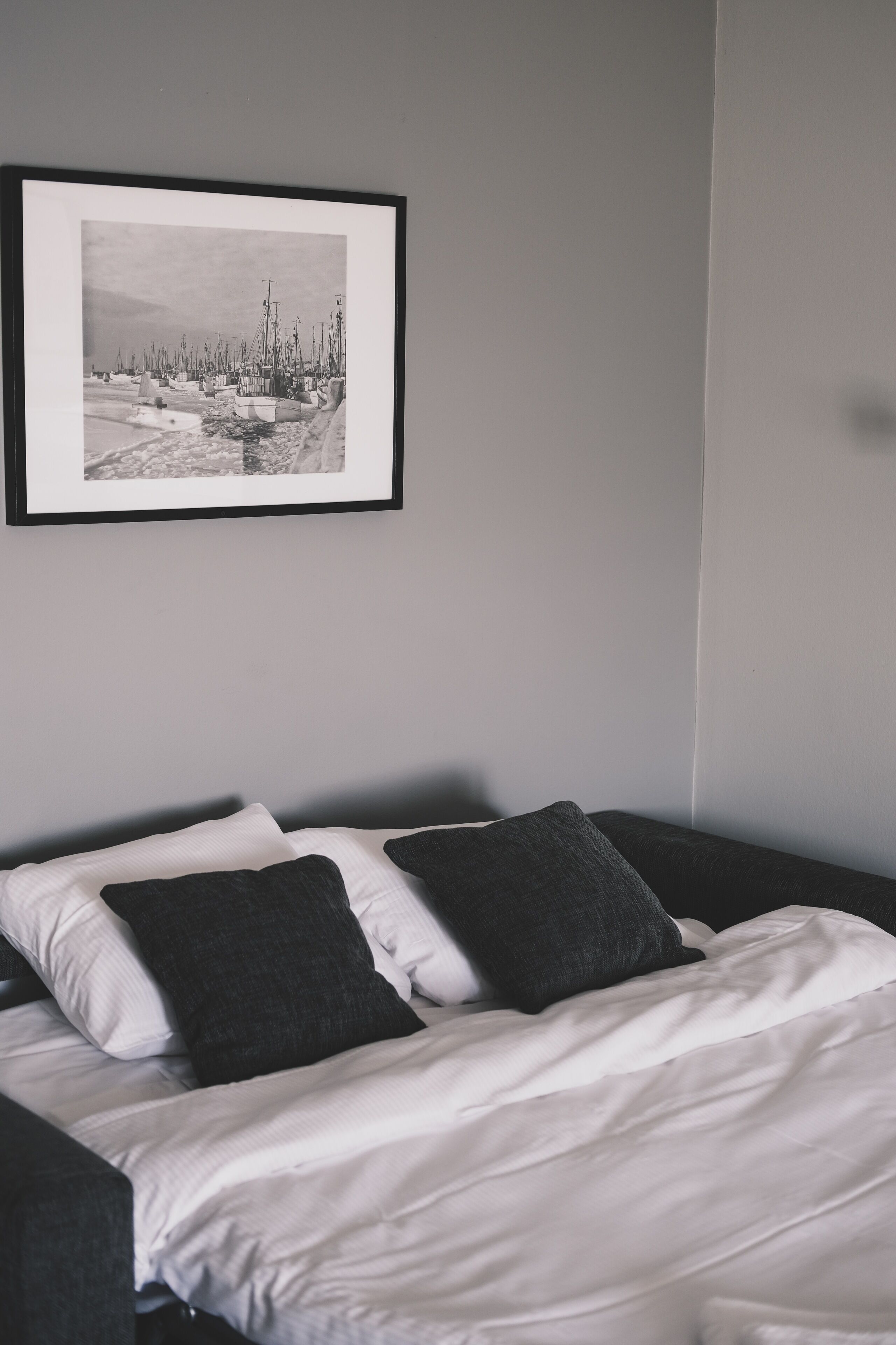 Una cama bien tendida con almohadas blancas y oscuras, una fotografía en blanco y negro enmarcada en la pared y una pared lisa.