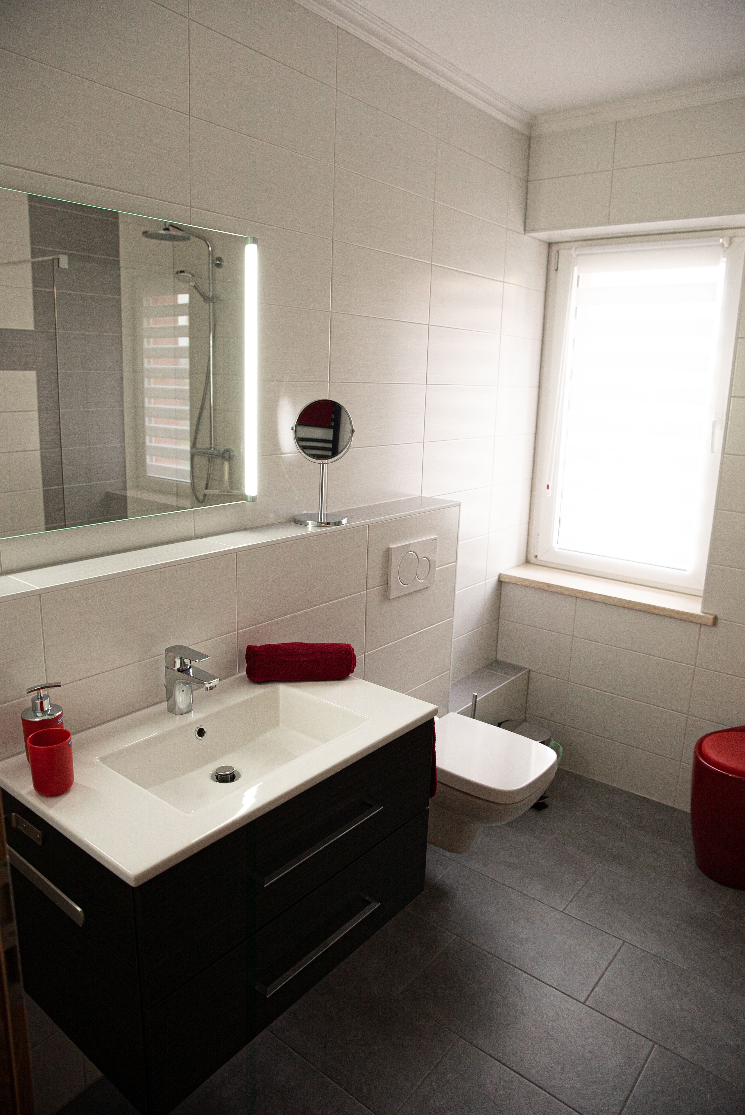 Un baño moderno con un lavabo blanco, un mueble negro, una toalla roja y una ventana con persianas.