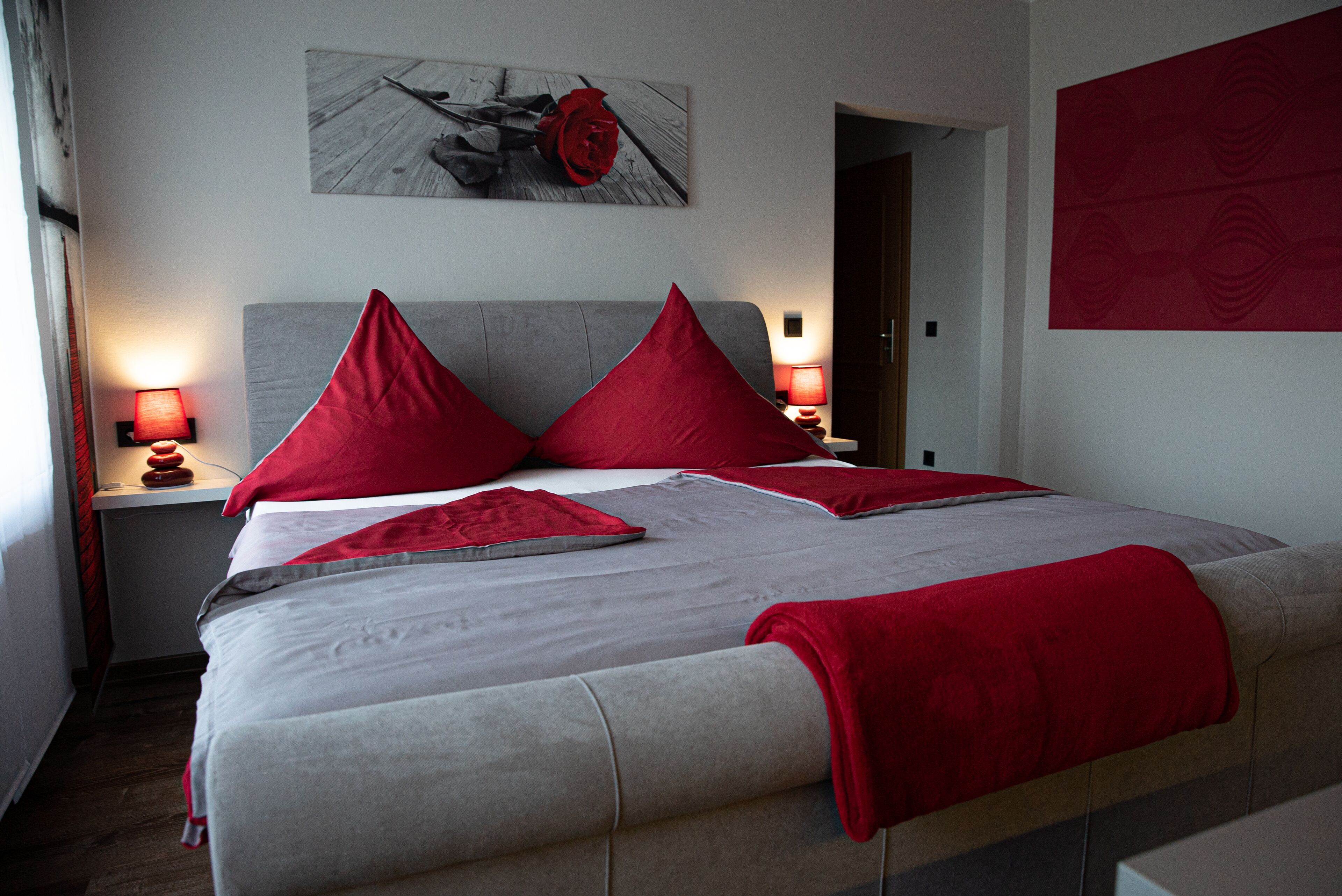 Un lit bien fait, avec des oreillers rouges et une tête de lit grise, une œuvre d’art murale aux couleurs rouge et blanche au-dessus, et un mur à motifs rouges sur le côté droit.
