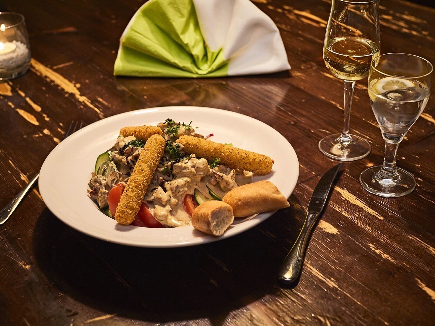 A plate of food with breadsticks, a salad, and a roll, accompanied by two glasses of white wine on a wooden table.