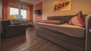 A hotel room with a sofa bed, a TV mounted on the wall, a painting above the bed, and a window with curtains. - Restaurant & Pension Rüssel - Pub (Sangerhausen)