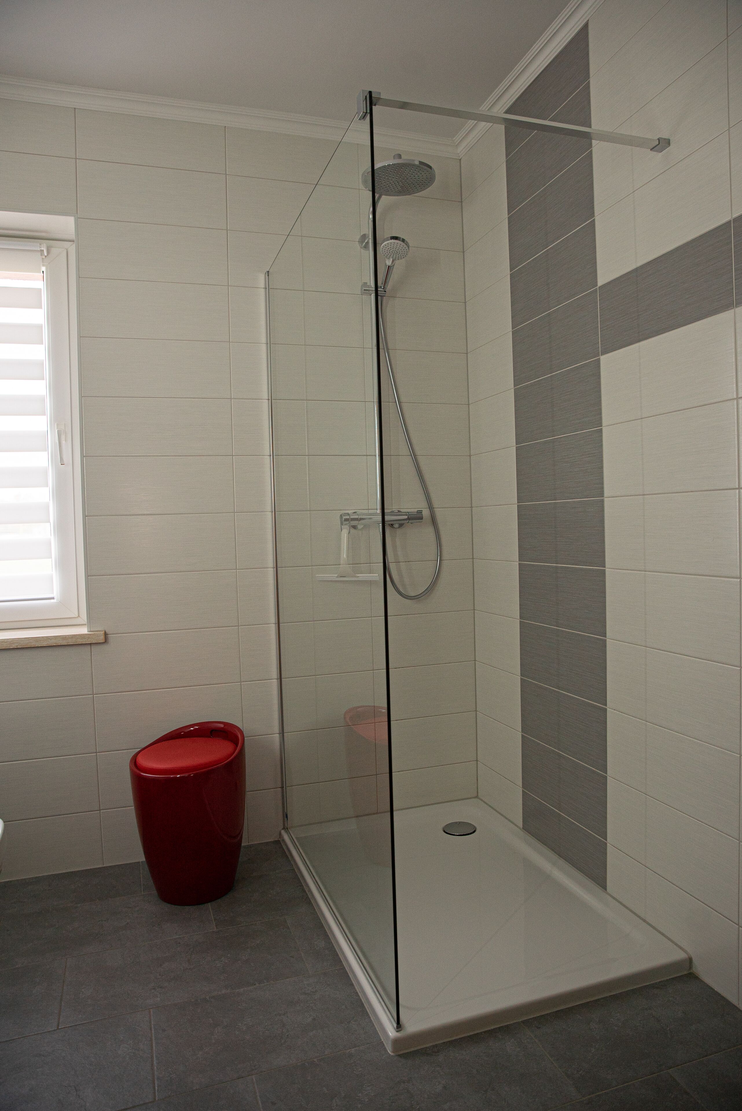 Una ducha con puerta de cristal, un cubo de basura rojo y una ventana con persianas.
