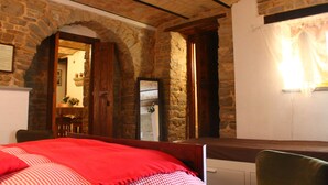 A room with a stone wall, a bed with a red blanket, a wooden chair, and a white door. - B&B Il Tomolo (Agnone)