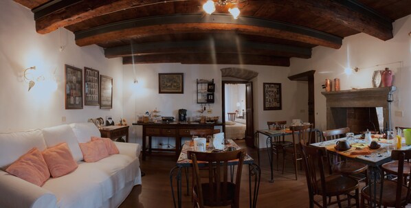 A cozy living room with a white sofa, wooden dining table, and chairs. There is a fireplace, a kitchen area, and various framed pictures on the walls. - B&B Il Tomolo (Agnone)