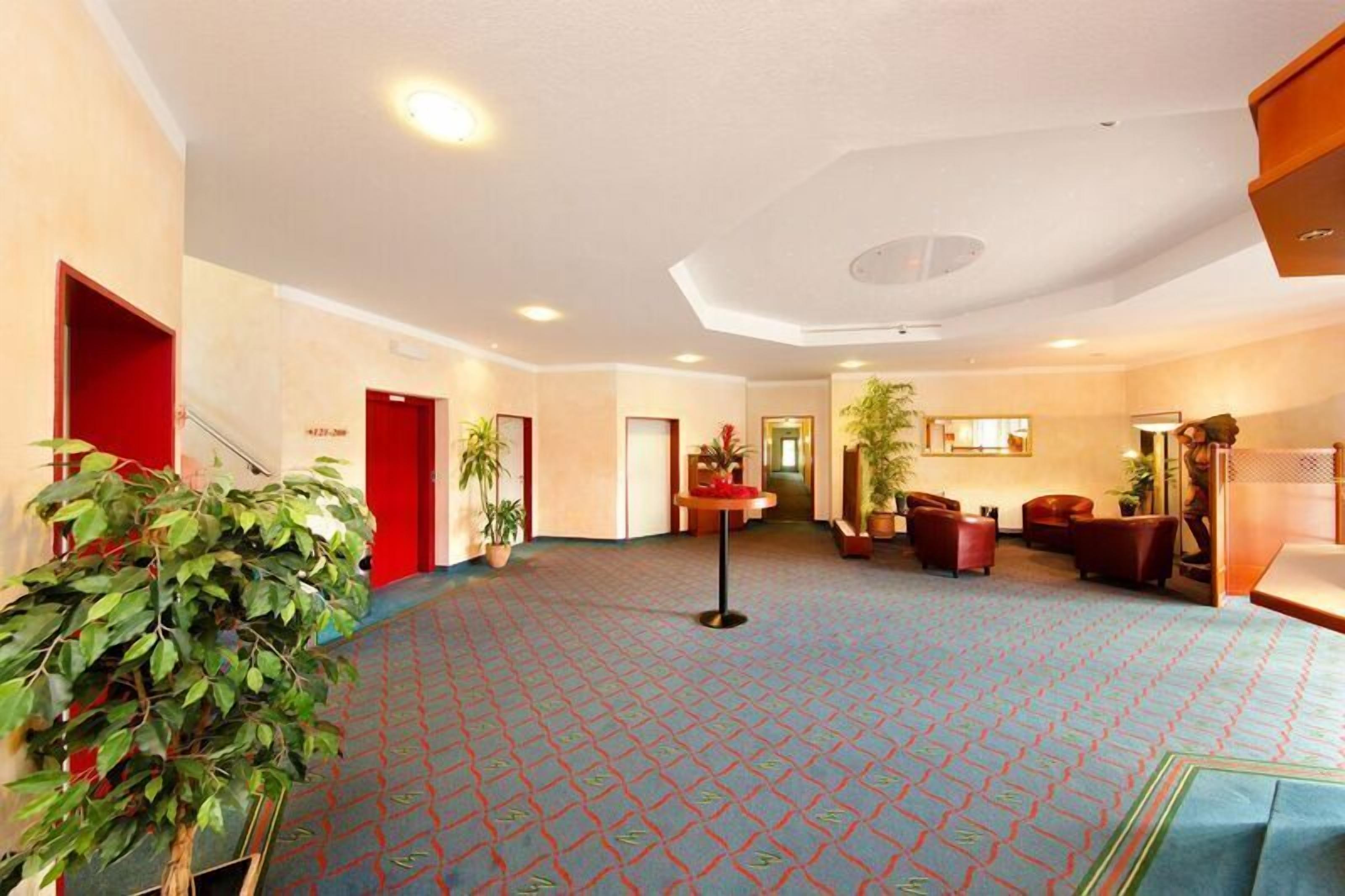 Eine großzügige Lobby mit einem gemusterten Teppich, roten Türen und einer Sitzecke mit Ledersesseln.