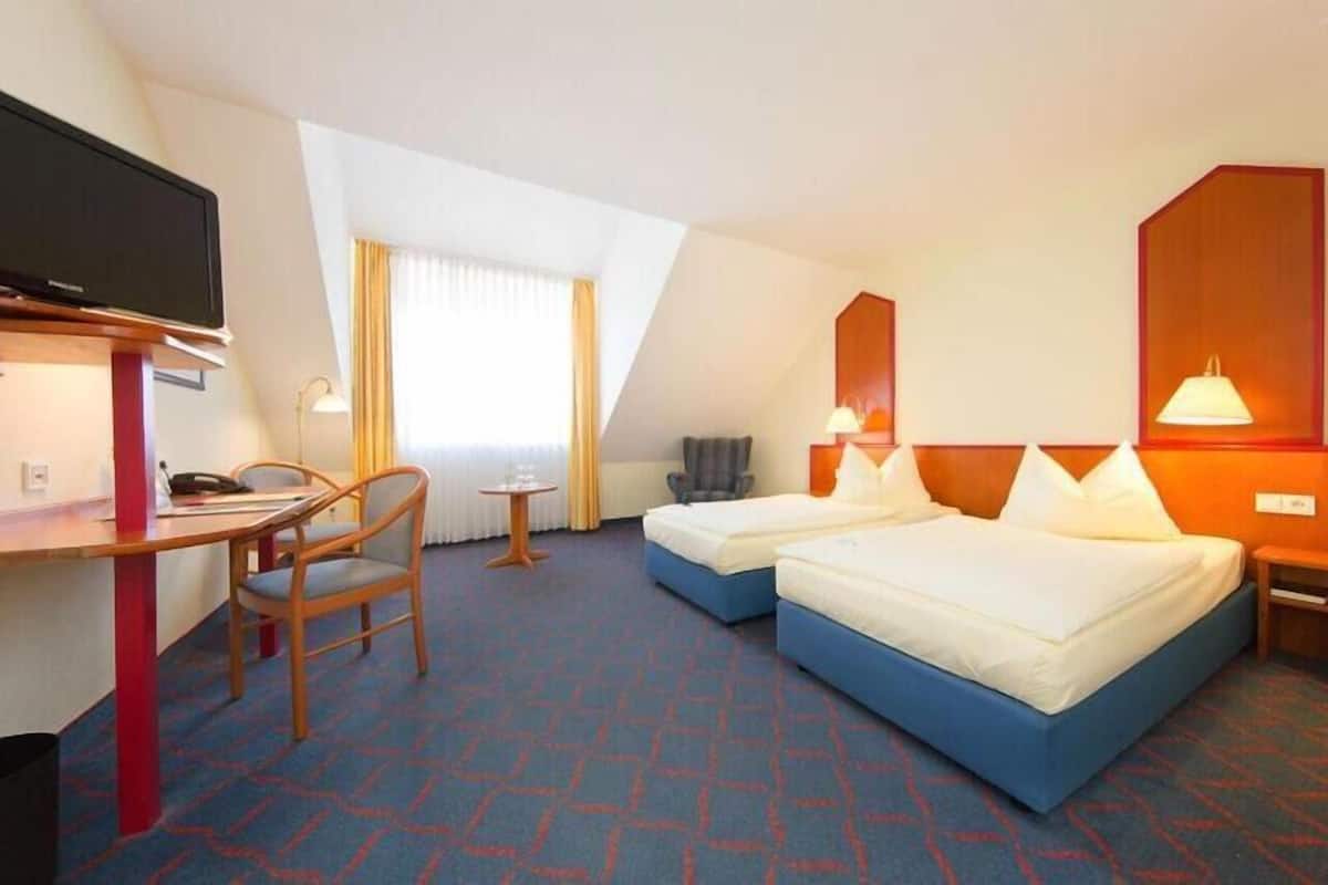 Ein Hotelzimmer mit zwei Betten, einem Schreibtisch mit Stuhl, einem Fernseher und einem Fenster mit Vorhängen.