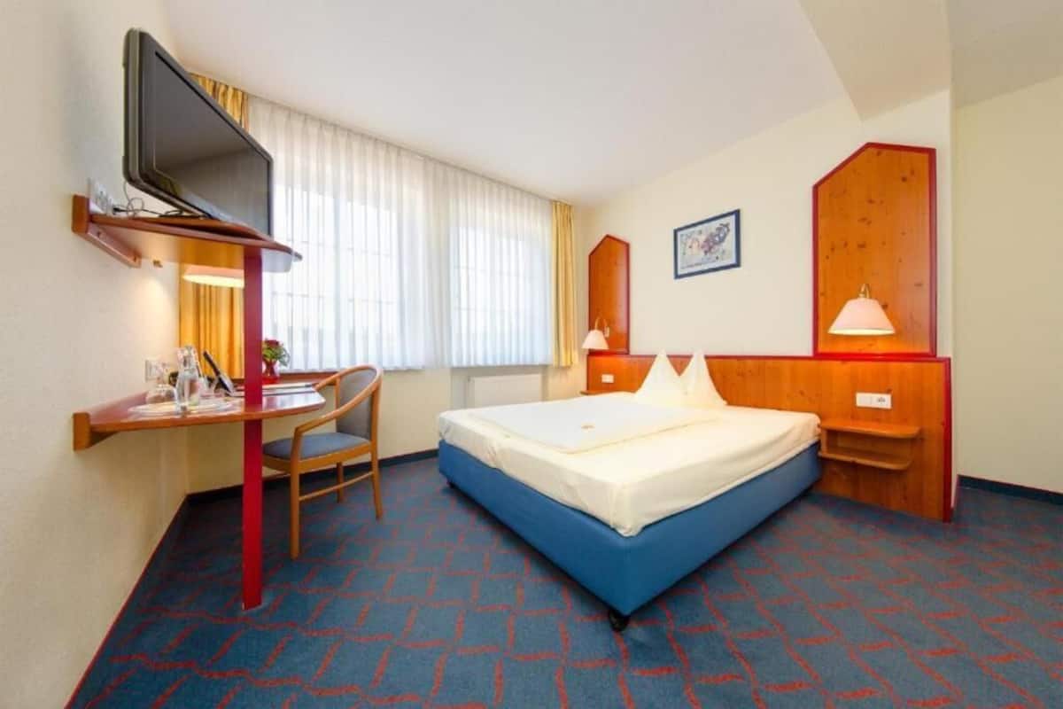 Ein Hotelzimmer mit Bett, Schreibtisch mit Stuhl, Wandfernseher und einem Vorhangfenster.