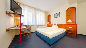 A hotel room with a bed, a desk with a chair, a television mounted on the wall, and a window with curtains.