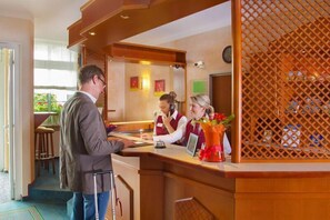 A hotel reception area with a wooden reception desk, a waiting area with a bar, and a view of the outdoors. - Montana Hotel Kassel-Süd (Guxhagen)