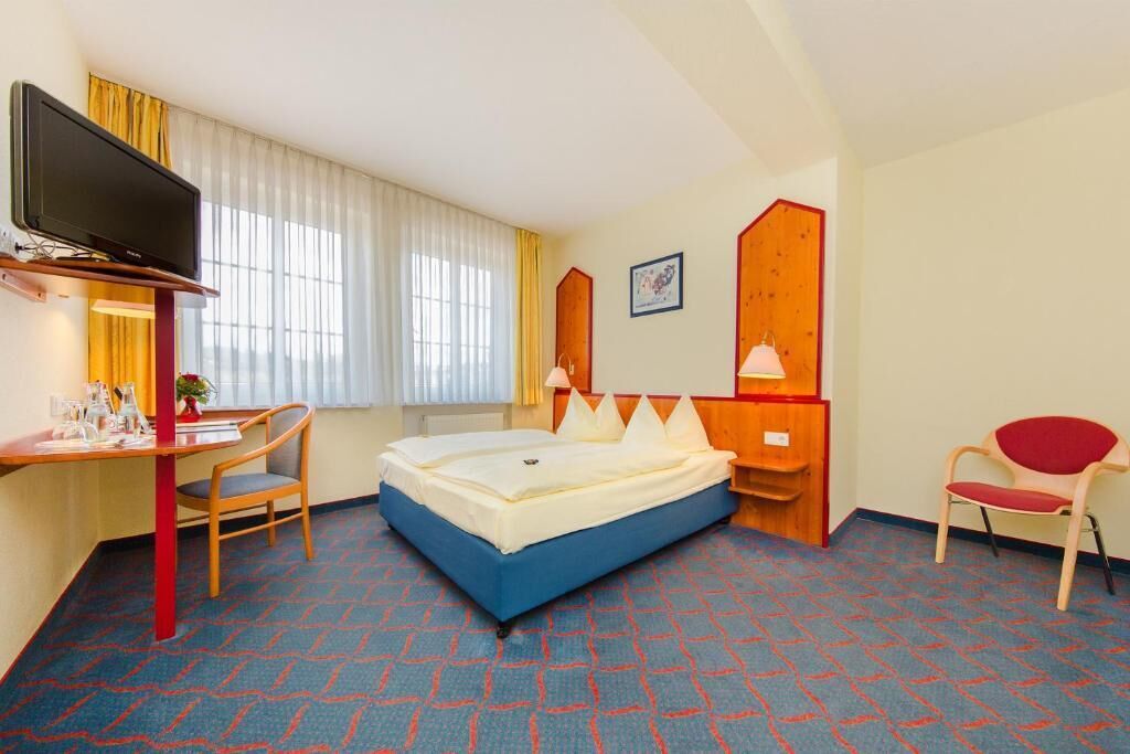 Ein Hotelzimmer mit einem Bett, einem Schreibtisch mit Stuhl, einem Fernseher und einem Fenster mit Vorhängen.