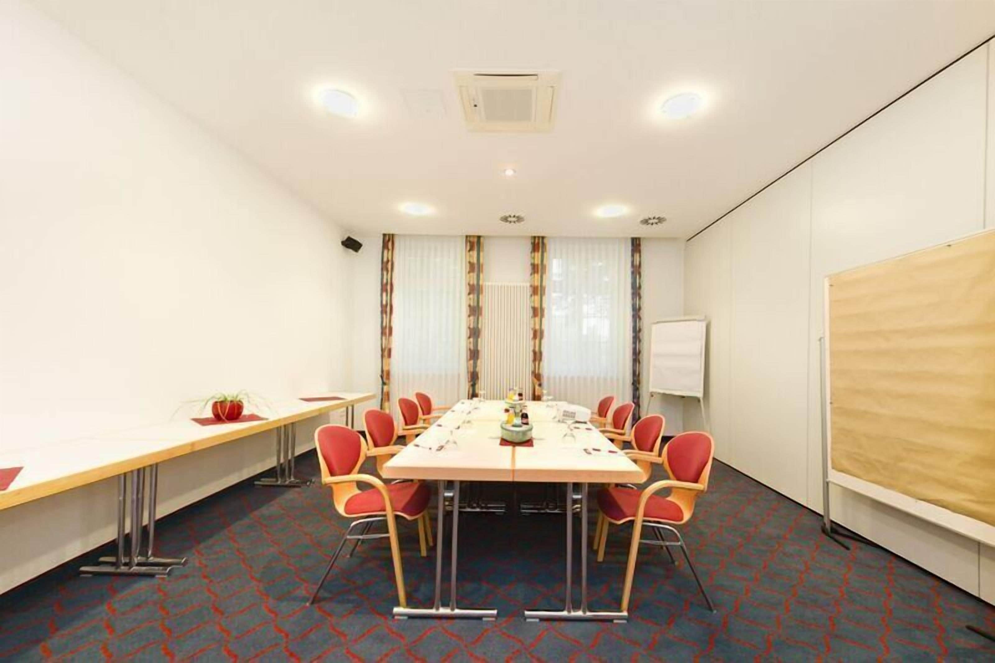 Une salle de conférence avec une longue table, des chaises rouges, un tableau blanc et un écran de projection.
