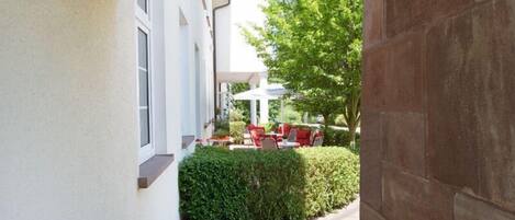 A covered outdoor seating area with red chairs and a table, a hedge, and a building with a white facade.