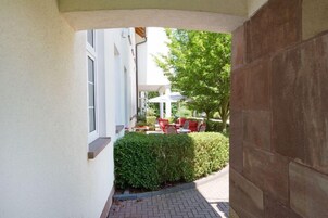 A covered outdoor seating area with red chairs and a table, a hedge, and a building with a white facade.