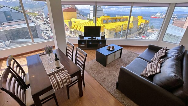 Ein modernes Wohnzimmer mit Essbereich, großen Fenstern mit Blick auf Stadt und Berge, einem Flachbildfernseher und einer Couch mit Kissen.