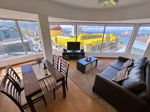 A modern living room with a dining area, large windows with a view of a cityscape and mountains, a flat-screen TV, and a sofa with cushions. - Patagonia Home Center (Ushuaia)