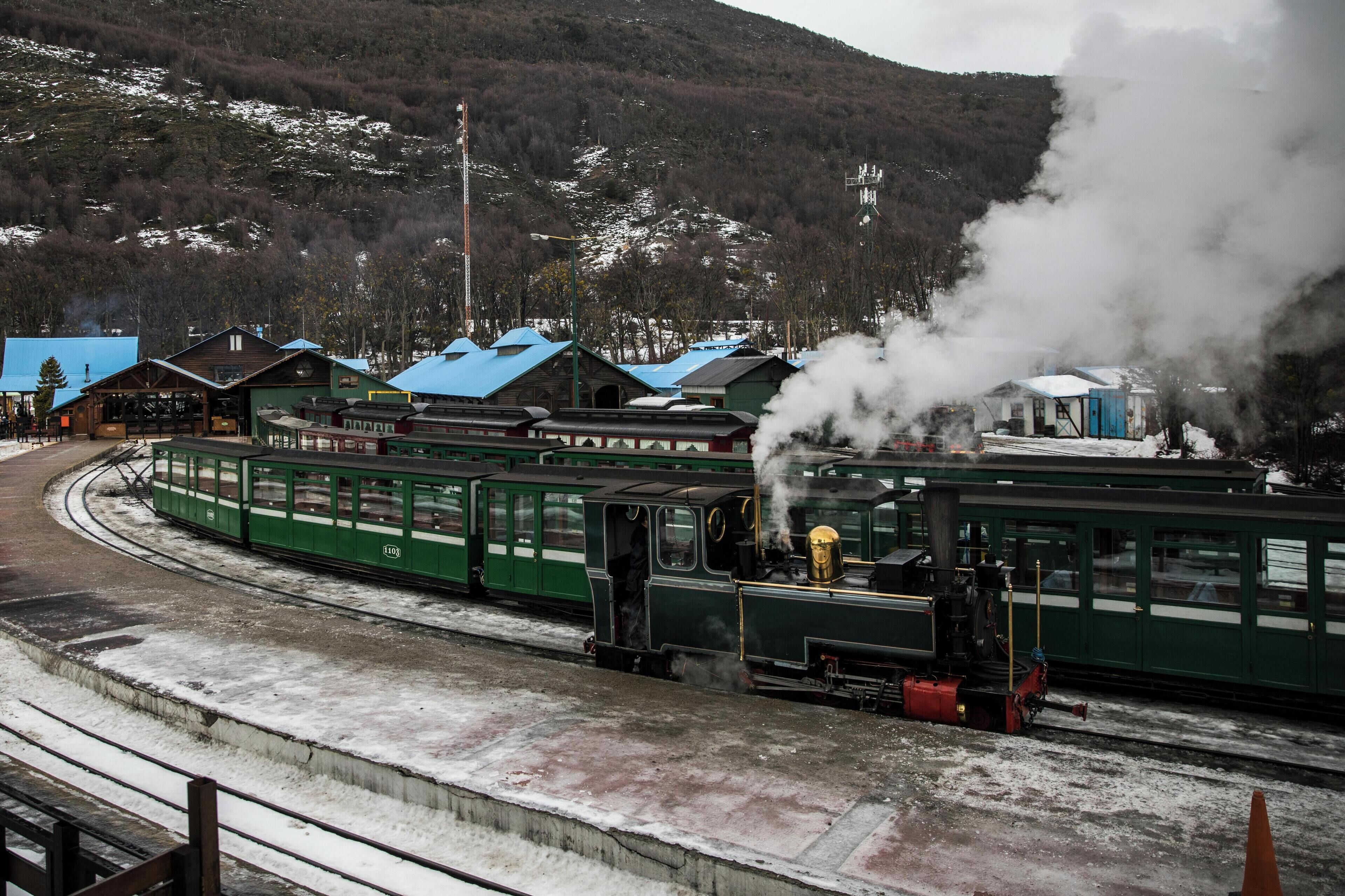 A green train with steam emitting from its engine, traveling on tracks with a mountainous backdrop.