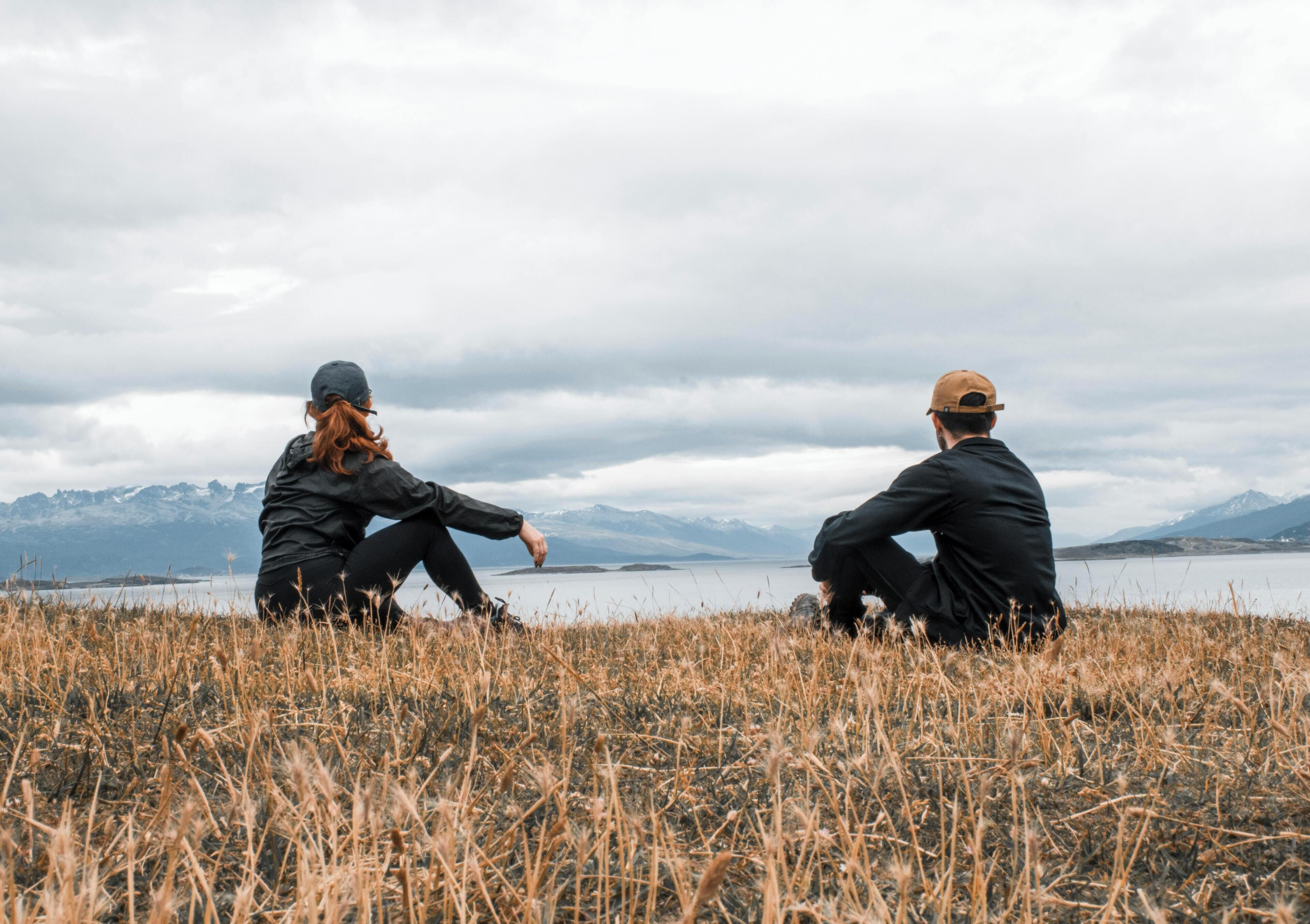 Deux personnes sont assises sur un champ d’herbe, avec un lac et des montagnes en arrière-plan.