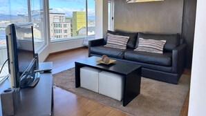 Ein modernes Wohnzimmer mit einer schwarzen Ledersofa, einem Couchtisch und einem Flachbildfernseher. Große Fenster bieten Blick auf Gebäude und Berge.