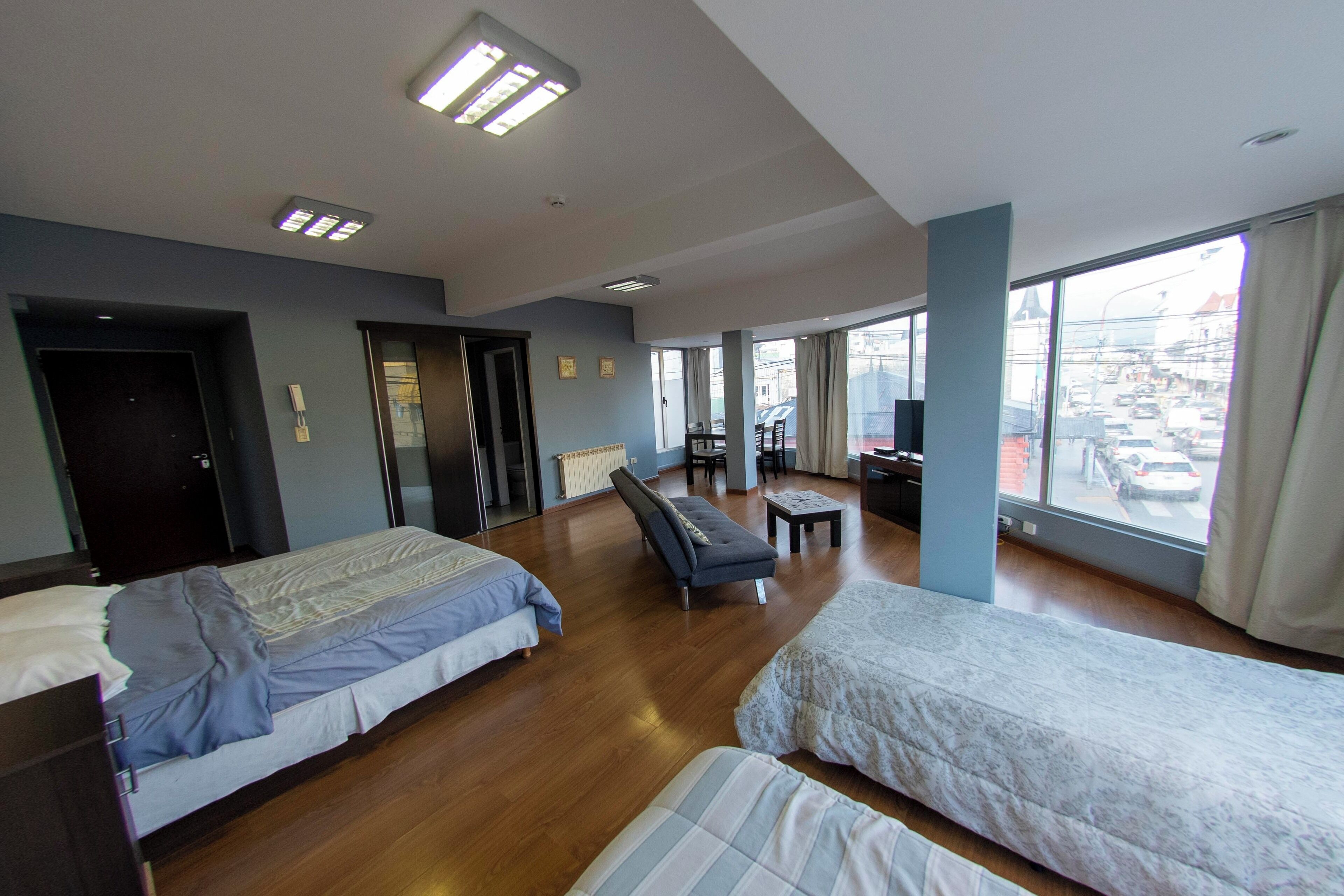 Habitación de hotel con dos camas, un sofá, una mesita y vista a la calle a través de amplias ventanas.