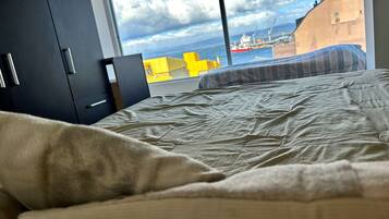 A bed with a view of a port and buildings through a window.