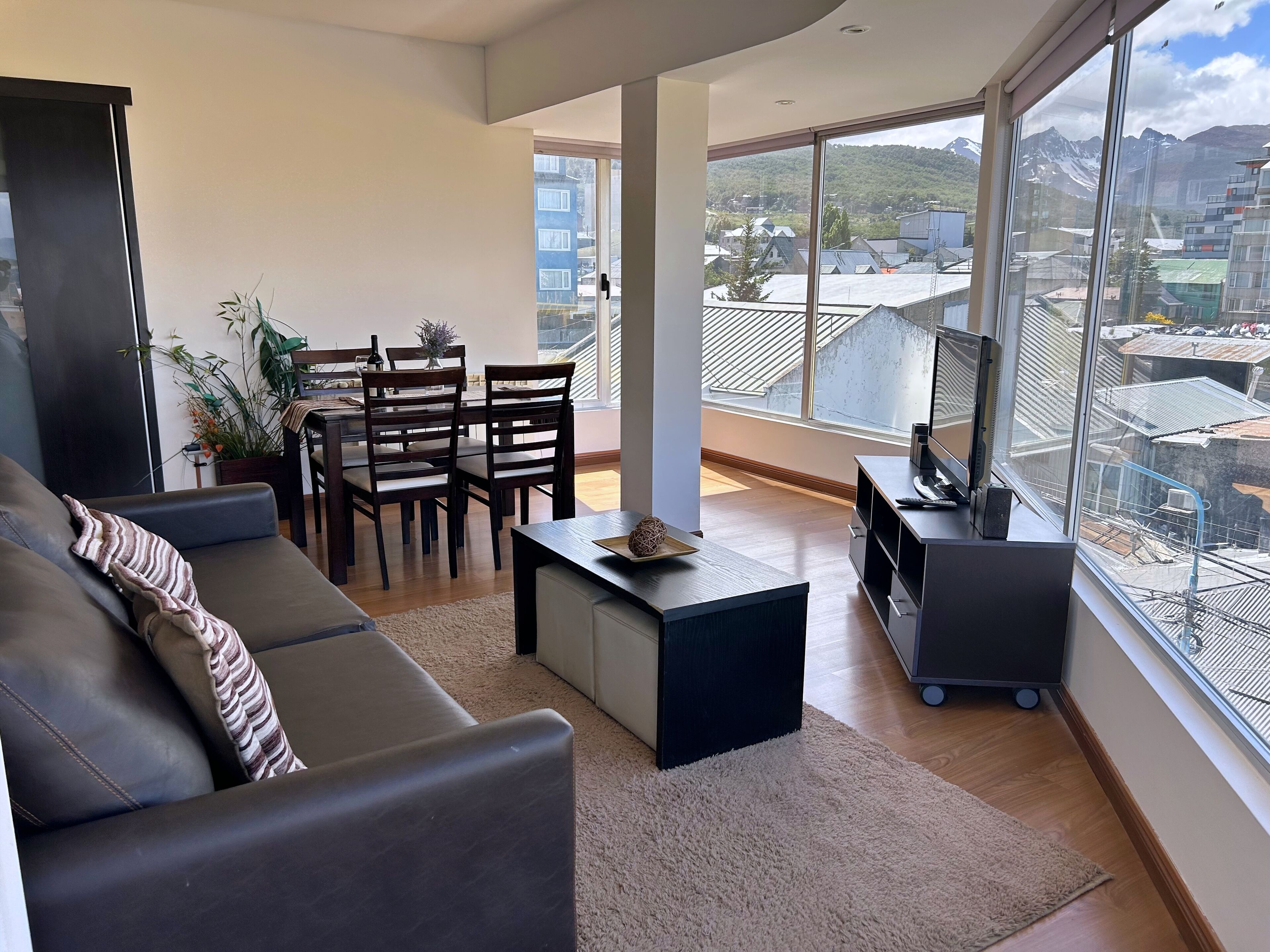 A modern living room with a sofa, a dining table with chairs, a coffee table, and a TV stand. Large windows offer a view of buildings and mountains.