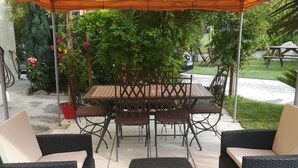 Pátio externo com mesa e cadeiras sob toldo, cercado por vegetação e um caminho de pedra.
