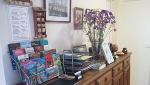 Uma receção com um armário de madeira, um quadro emoldurado, um vaso com flores roxas e um mural com diversos avisos.