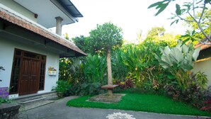 A courtyard with a central tree, a stone bench, and a pathway leading to a building with a tiled roof and a wooden door. - Akusara Guest House (Ubud)
