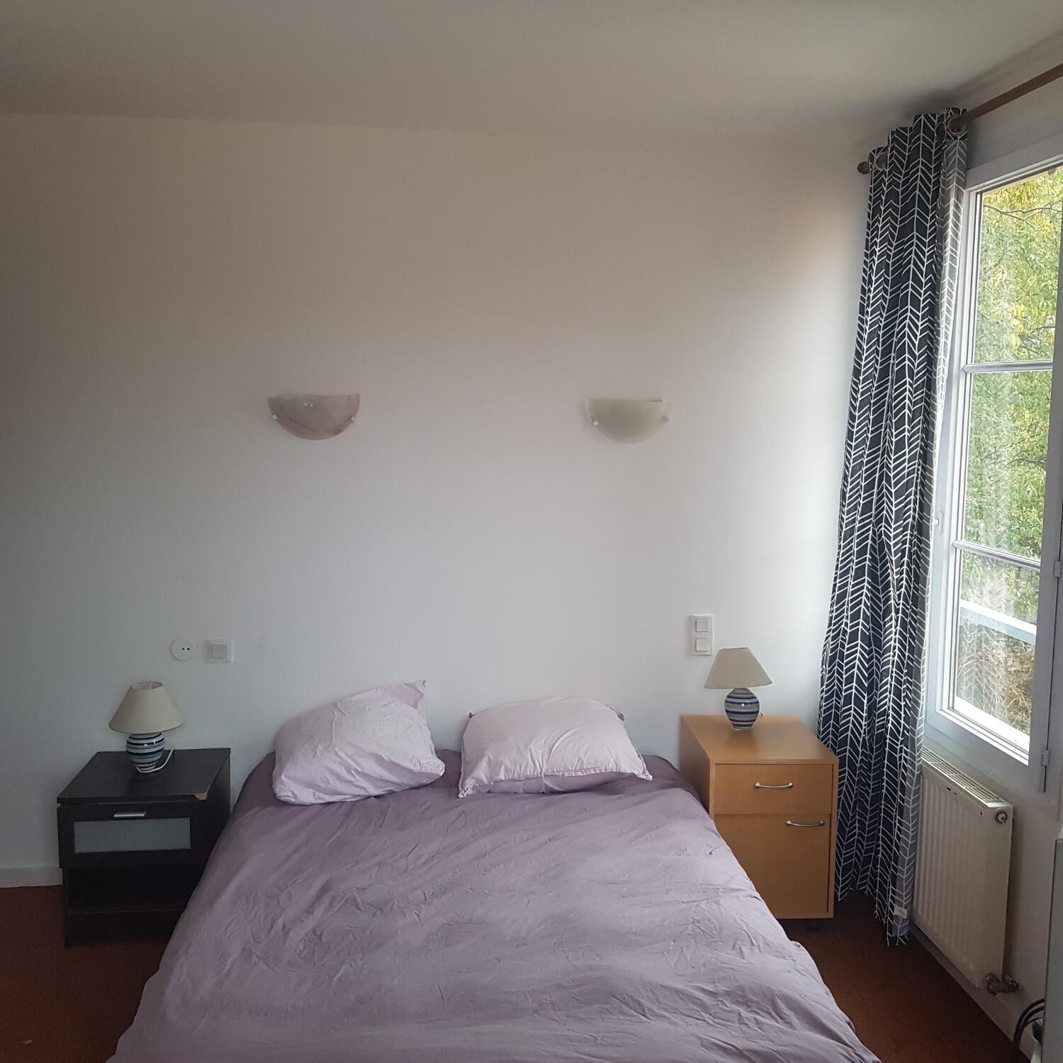 A bedroom with a bed, two bedside tables, a nightstand, a window with curtains, and a wall-mounted light fixture.