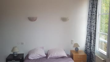 A bedroom with a bed, two bedside tables, a nightstand, a window with curtains, and a wall-mounted light fixture.