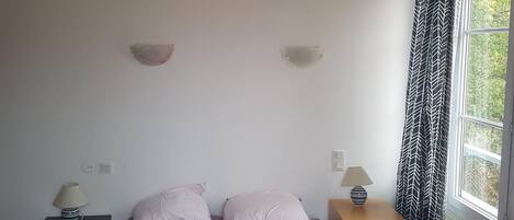 A bedroom with a bed, two bedside tables, a nightstand, a window with curtains, and a wall-mounted light fixture.