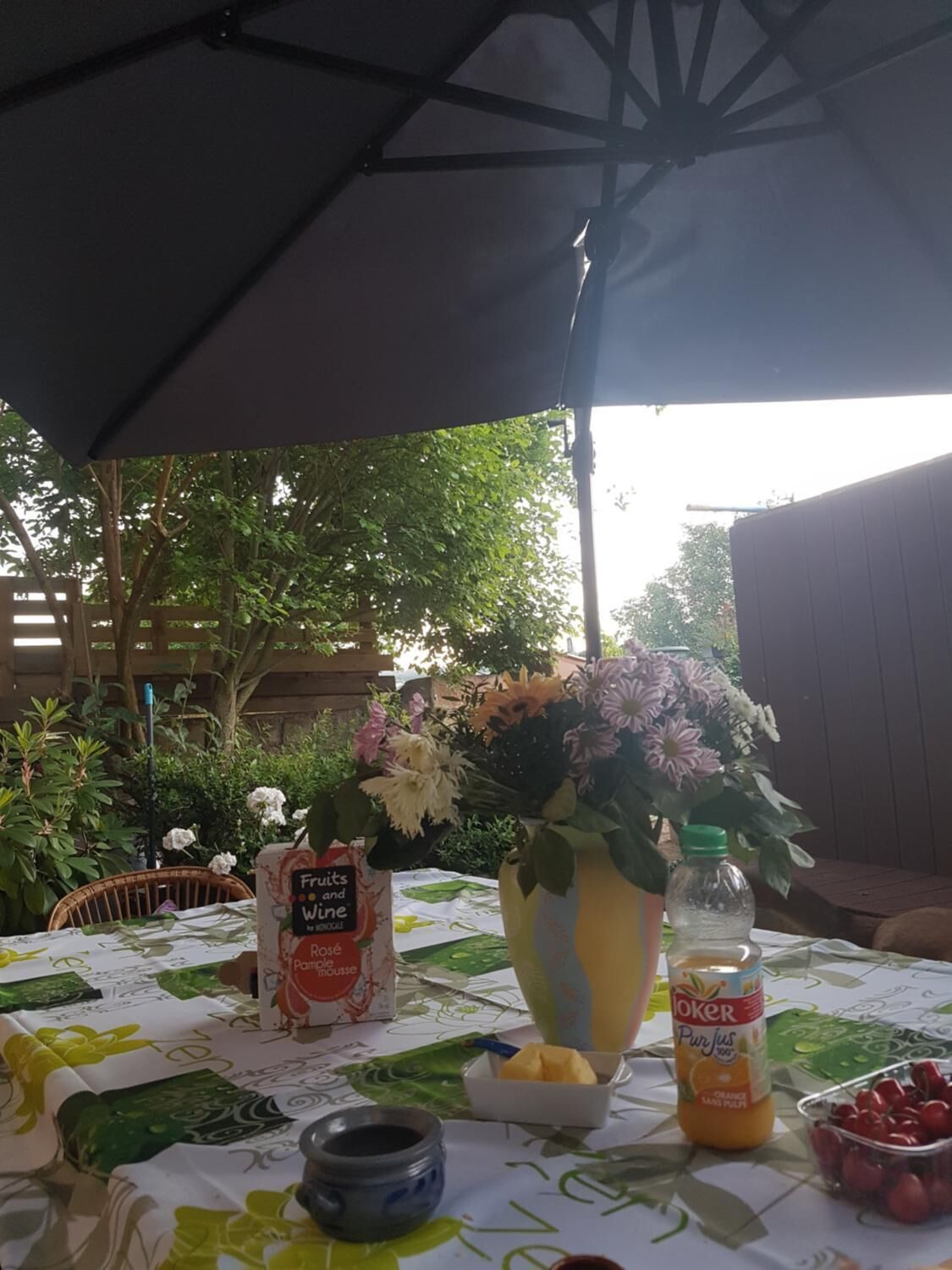 An outdoor table with a tablecloth, a vase of flowers, a bottle of juice, a bowl of cherries, and a container of jam.