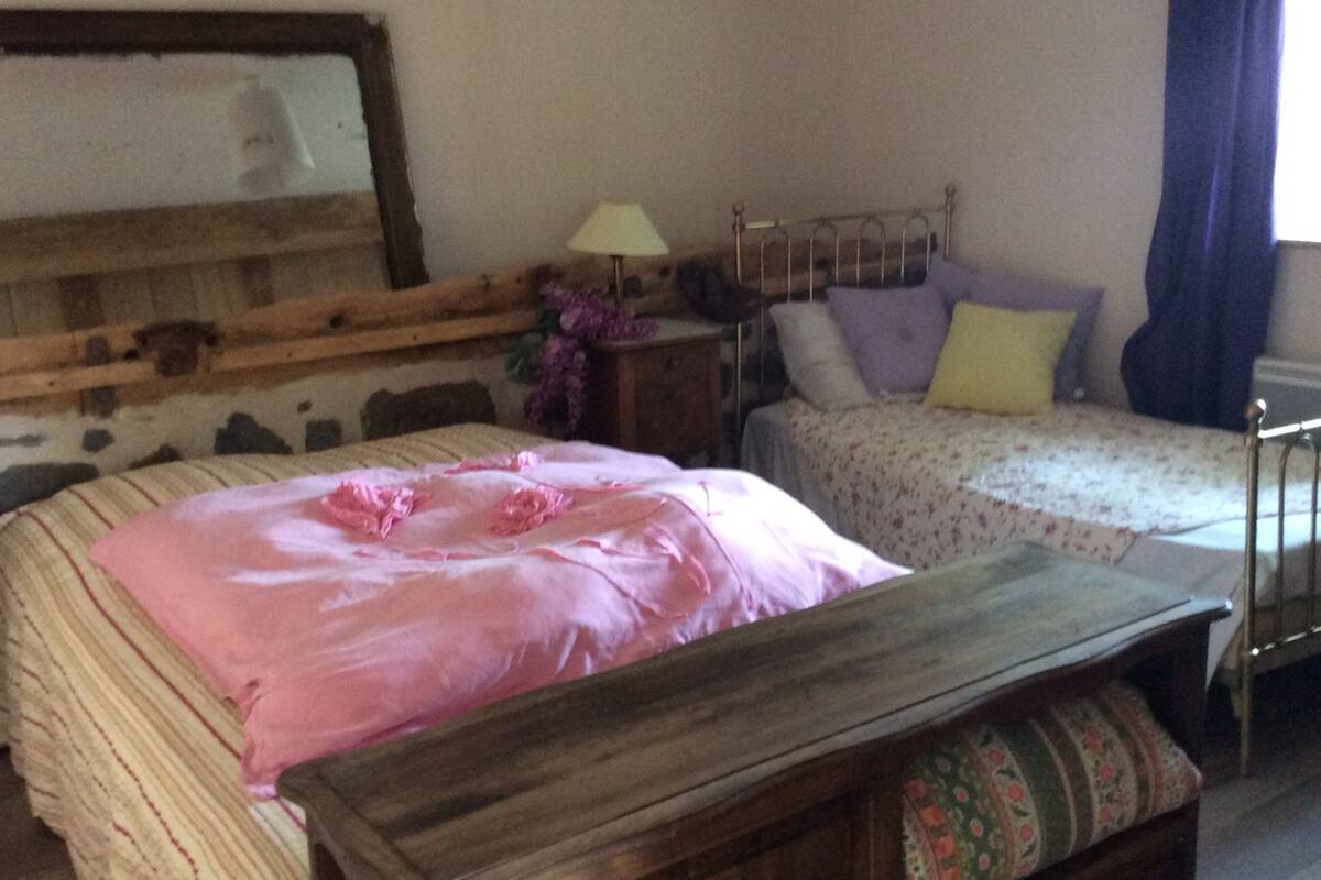 A bedroom with a large bed, a smaller bed, a wooden bench, a nightstand, a lamp, and a mirror on the wall.