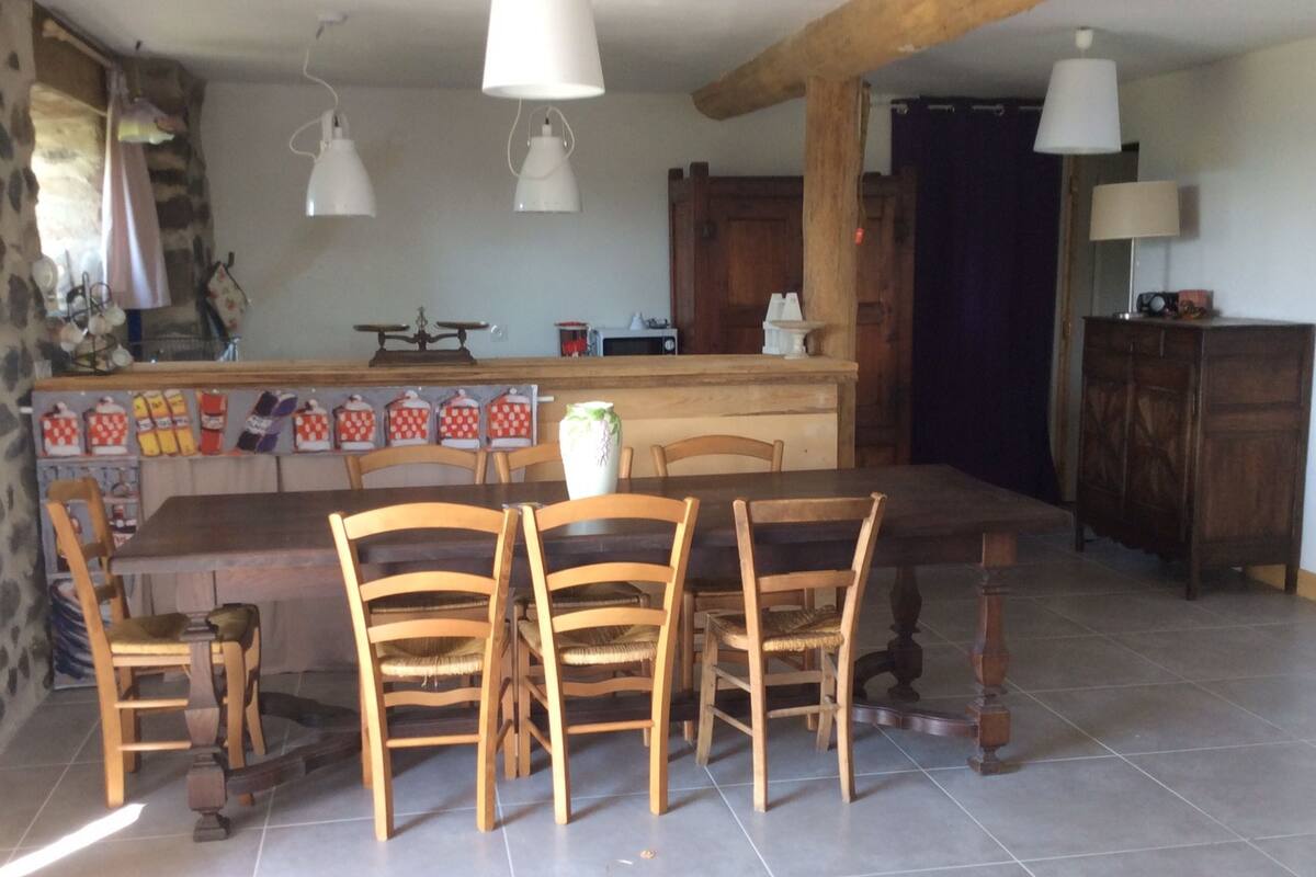 A dining area with a wooden table and chairs, a kitchen in the background, and hanging pendant lights.